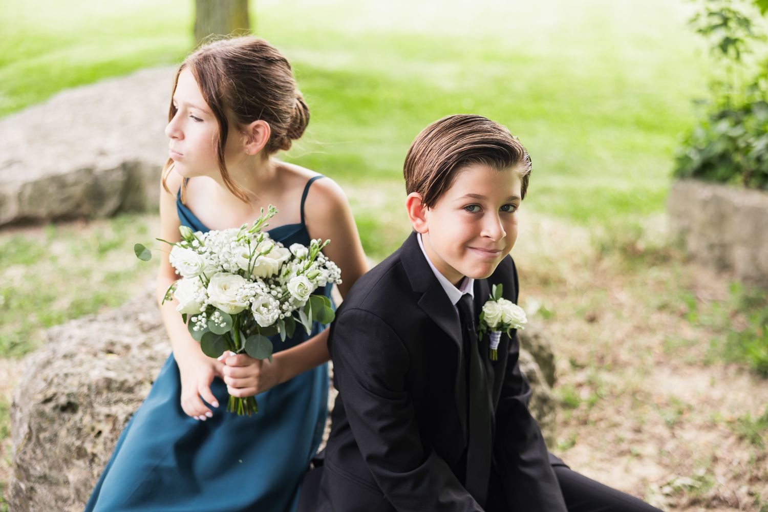 Young ring bearer in black suit and flower girl in teal dress holding white bouquet sit together at outdoor wedding.