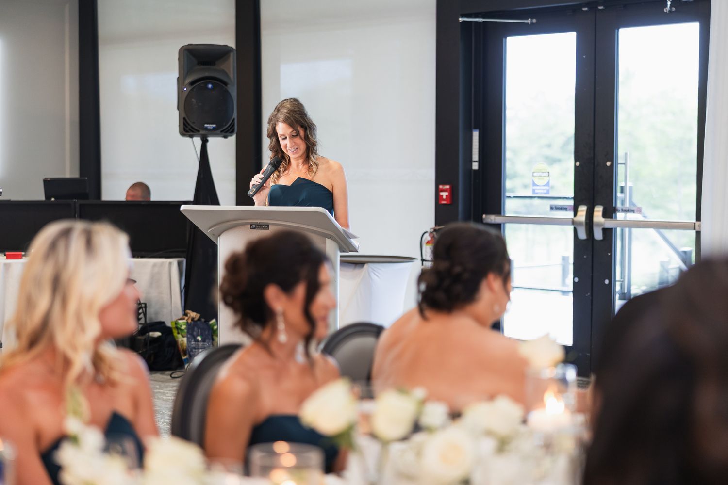 Woman in black dress delivers speech at podium during elegant wedding reception with floral centerpieces.