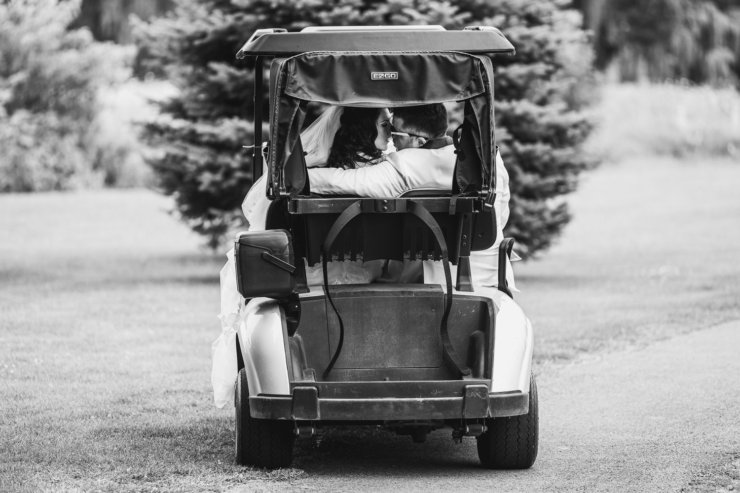 Black and white photo of a couple riding in a golf cart through a lush green park.