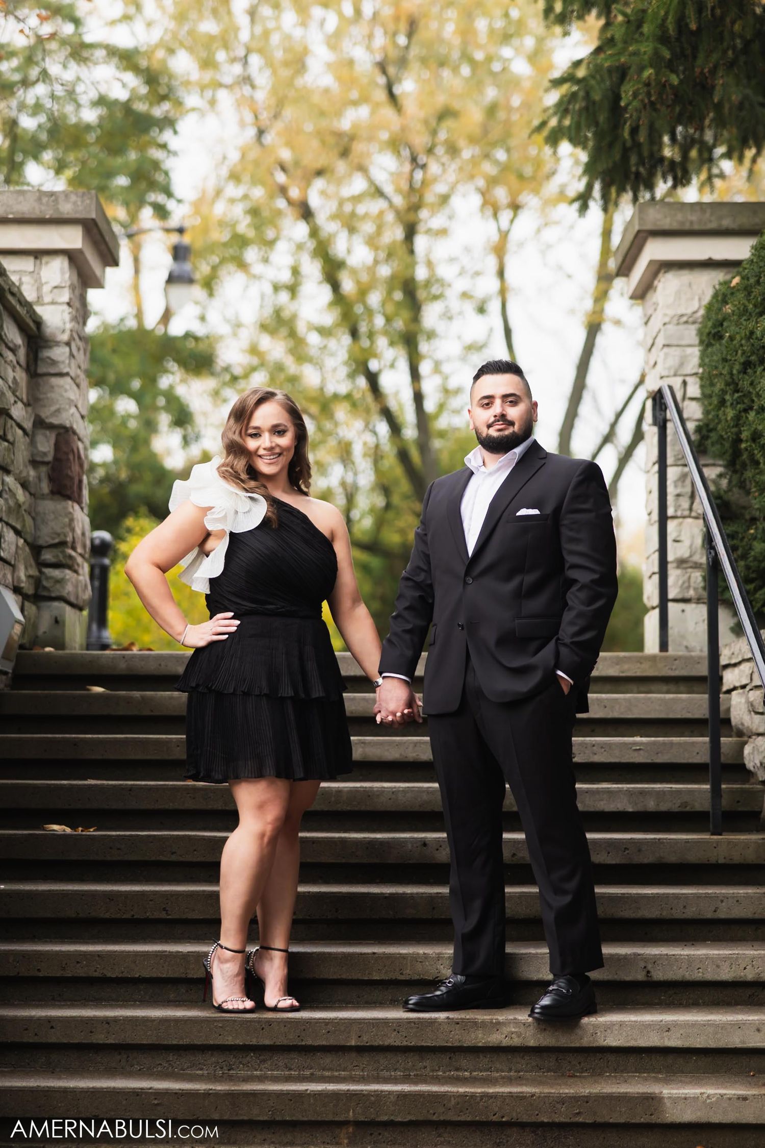 A couple poses on stone steps outdoors, with her in a black dress and him in a dark suit.