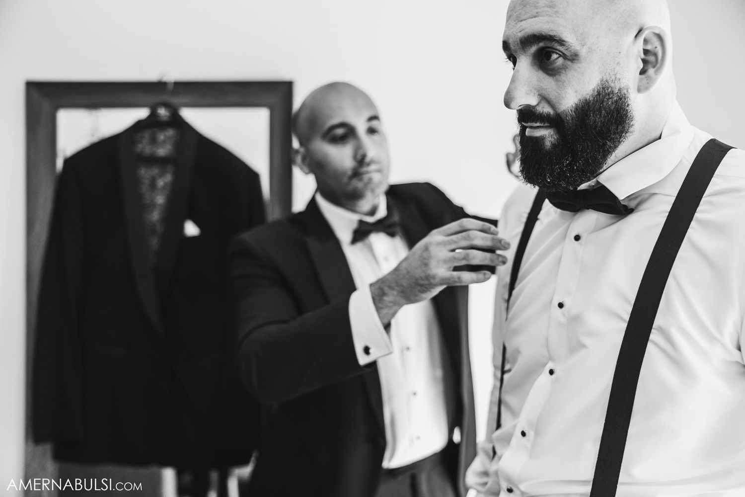 Two people in formalwear helping each other get dressed in front of mirror.