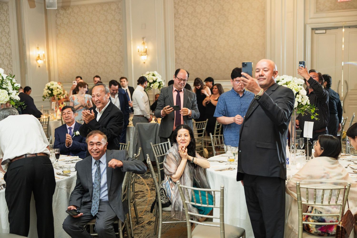 Wedding reception guests seated at decorated tables while someone speaks into a microphone.
