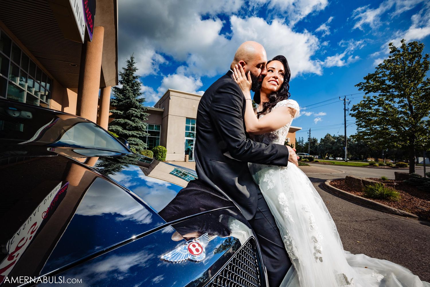 Newlyweds pose on car under blue sky during wedding photoshoot.