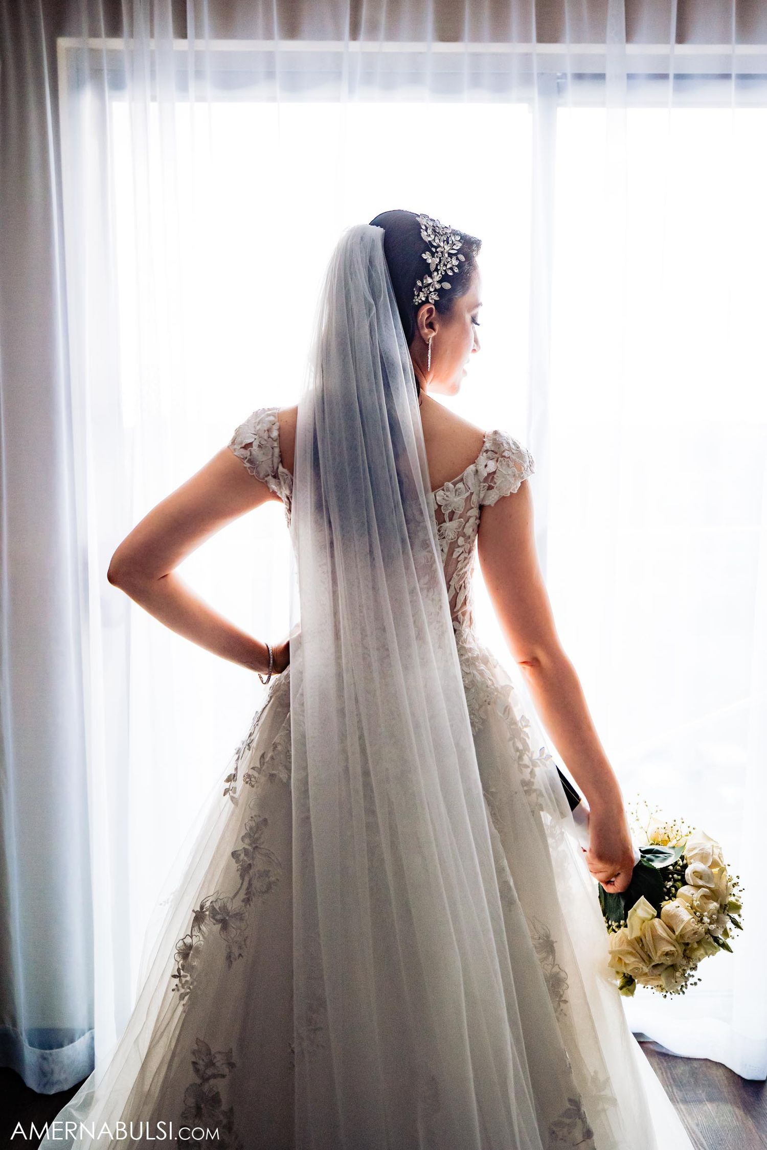 Elegant bride in lace dress and long veil poses by window holding white bouquet.