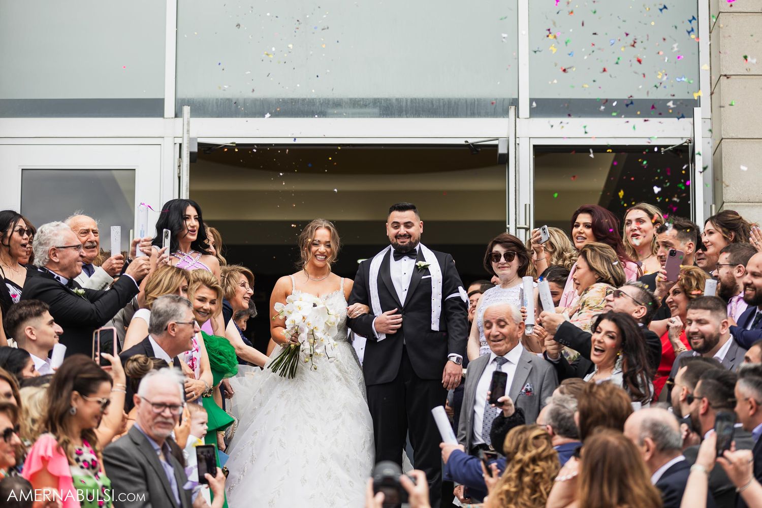 Wedding party and guests celebrate with confetti as couple stands on building steps.