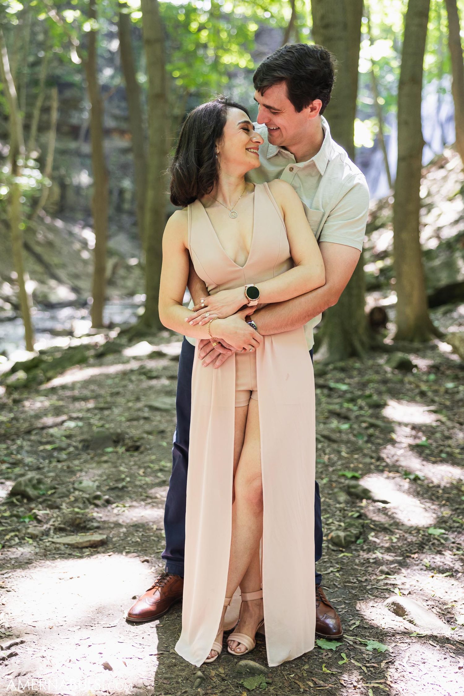 A couple embraces tenderly in a sun-dappled forest setting wearing complementary neutral-toned outfits.