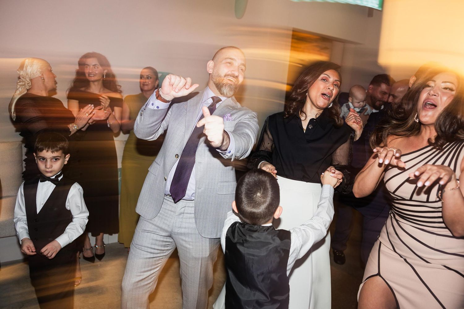 Party guests of all ages dance together at a lively indoor celebration.