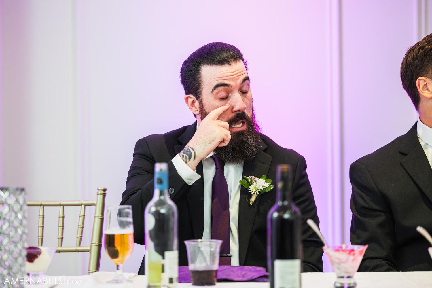 Wedding guest in black suit and boutonniere wiping his face at reception table.
