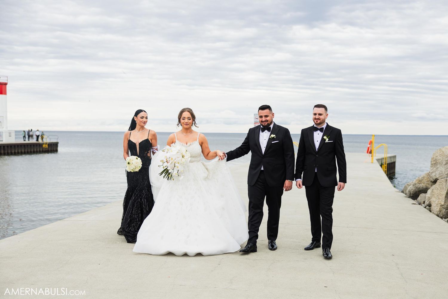 Wedding party walks along lighthouse pier under cloudy sky.