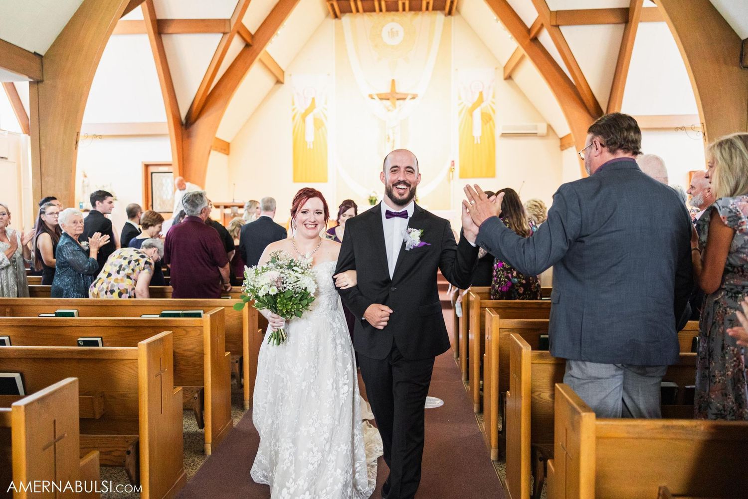 A wedding couple walks down the church aisle while guests look on from wooden pews.