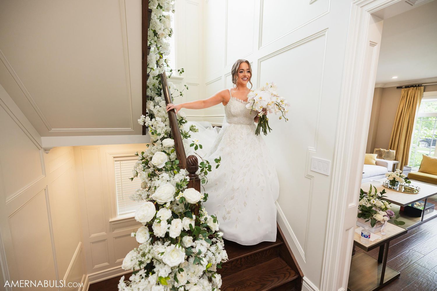 A bride in white poses elegantly on a staircase decorated with cascading white floral arrangements.