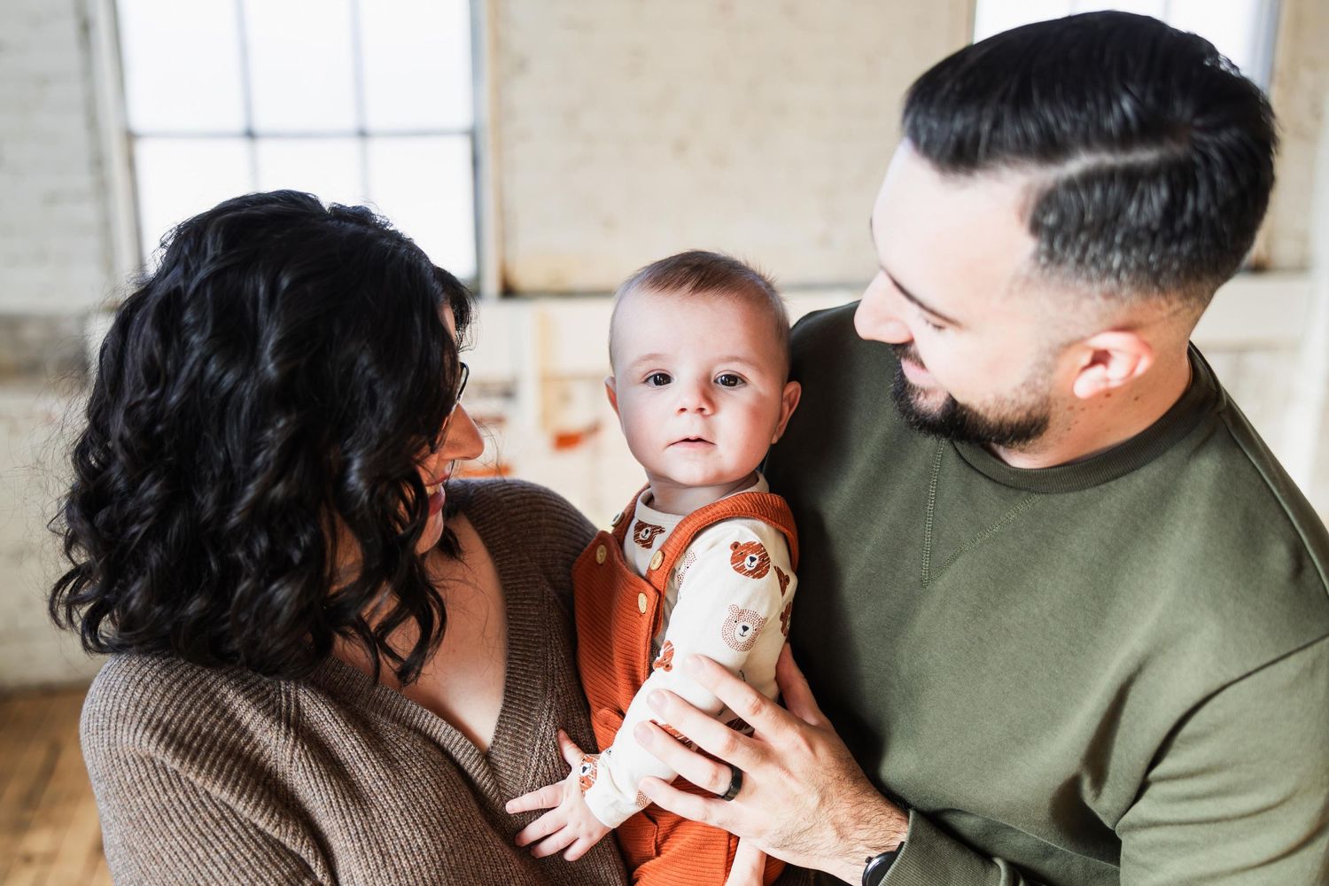 Loving parents holding their baby wearing an orange romper in a bright indoor setting.