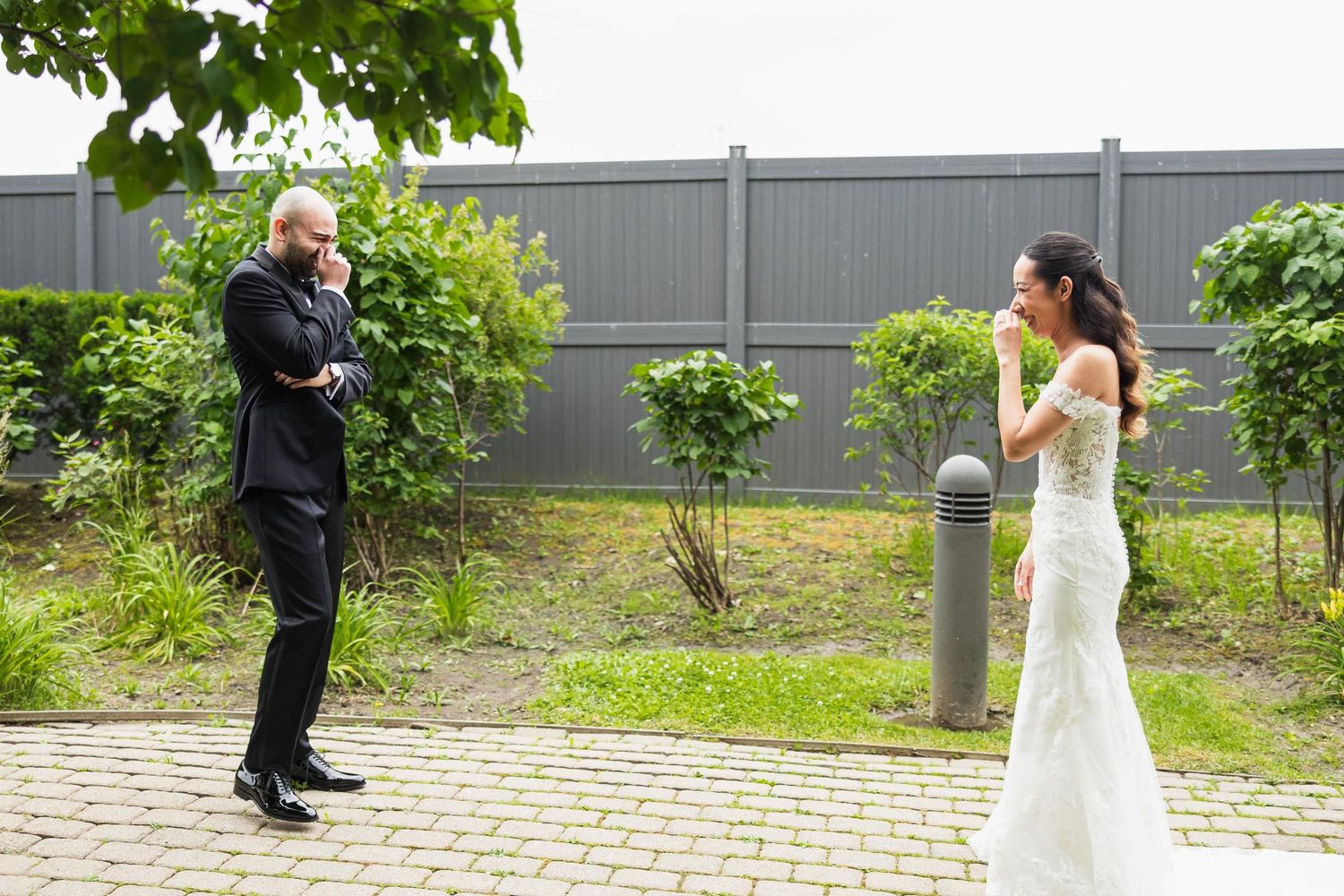 A couple sharing a candid moment outdoors on their wedding day with lush greenery and grey fencing in the background.
