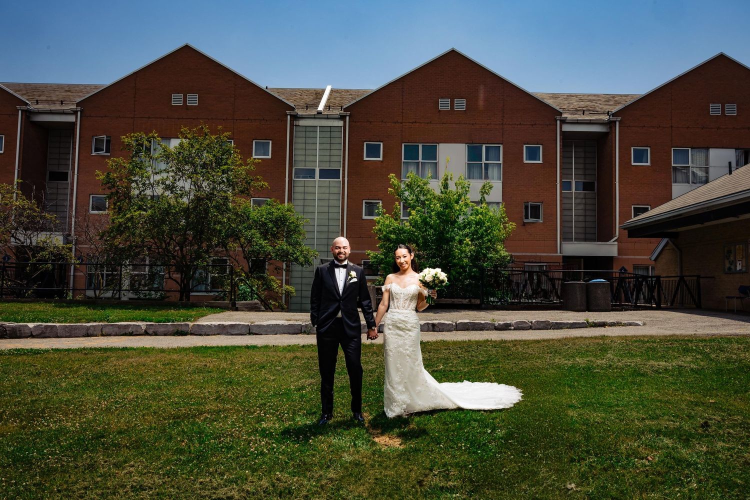 Wedding couple stands on lawn in front of red brick townhouse buildings.