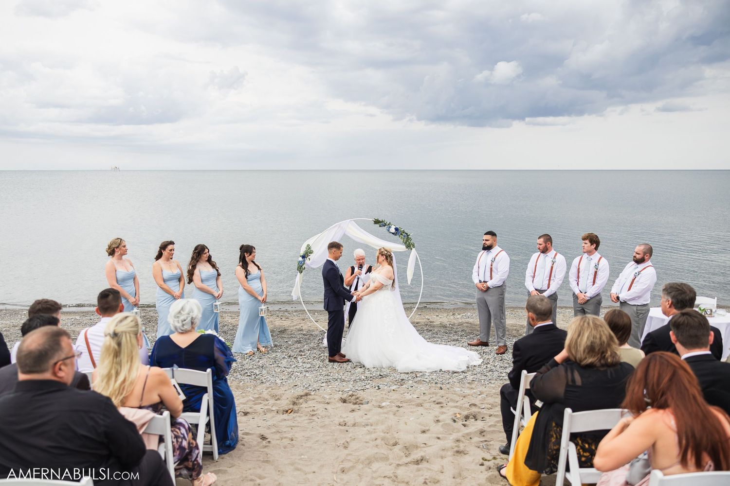 Wedding ceremony guests seated in white chairs along a scenic beach shoreline.