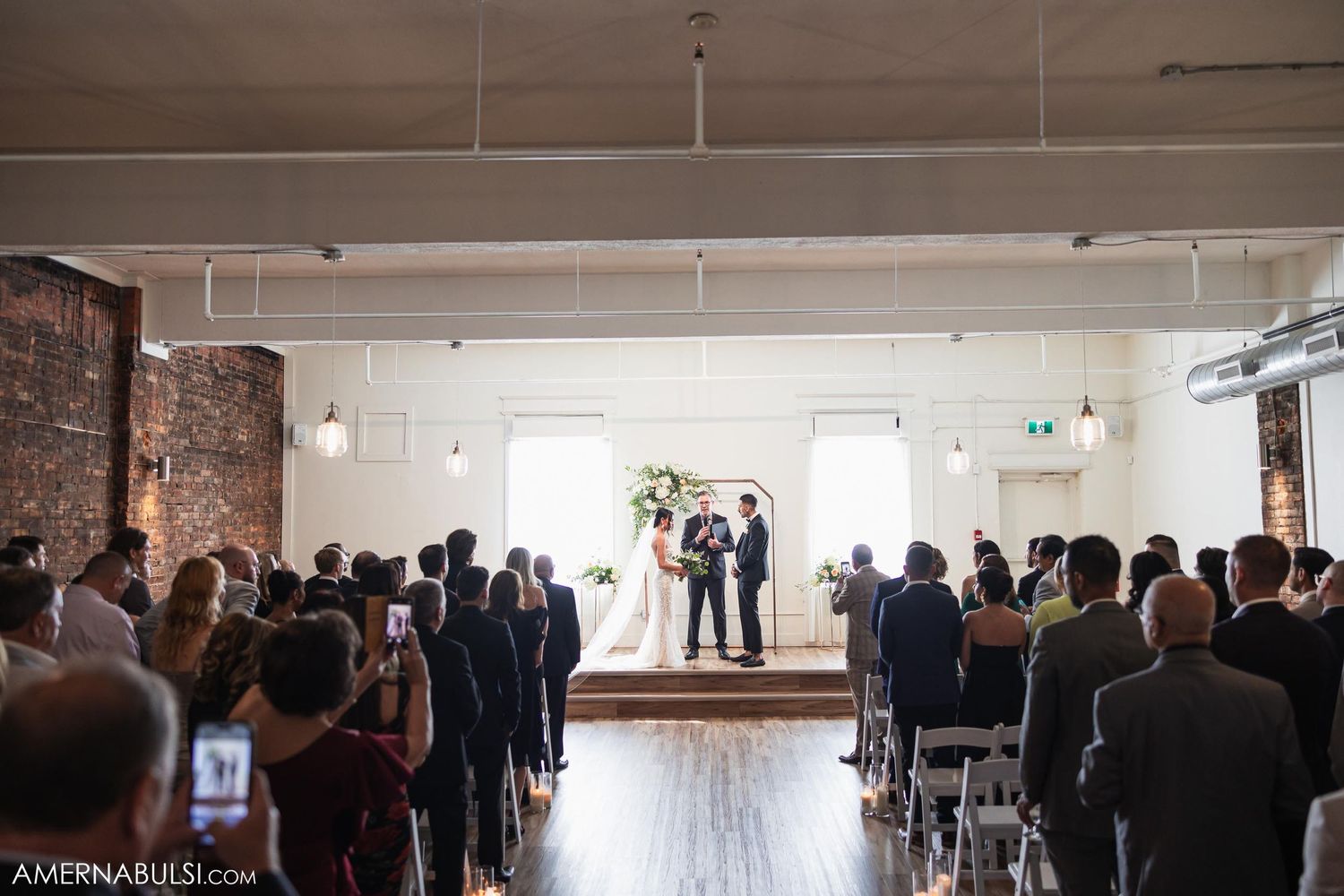 Wedding ceremony with guests seated in rows watching couple exchange vows in bright loft space.