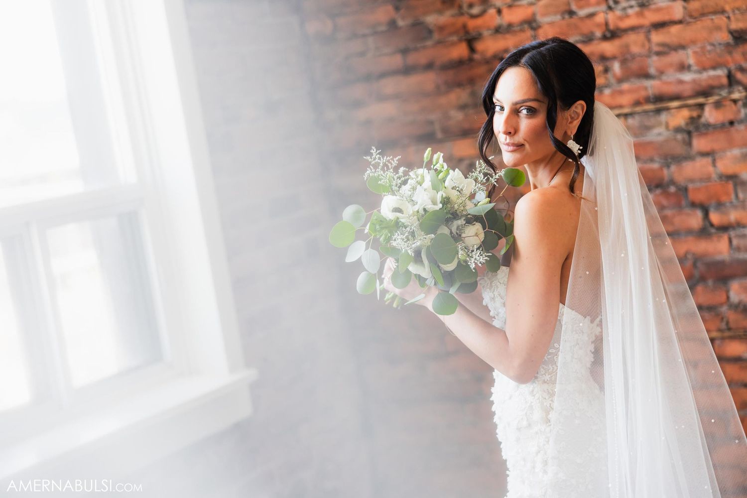 Bride in lace gown holds white bouquet against rustic brick wall with natural lighting.