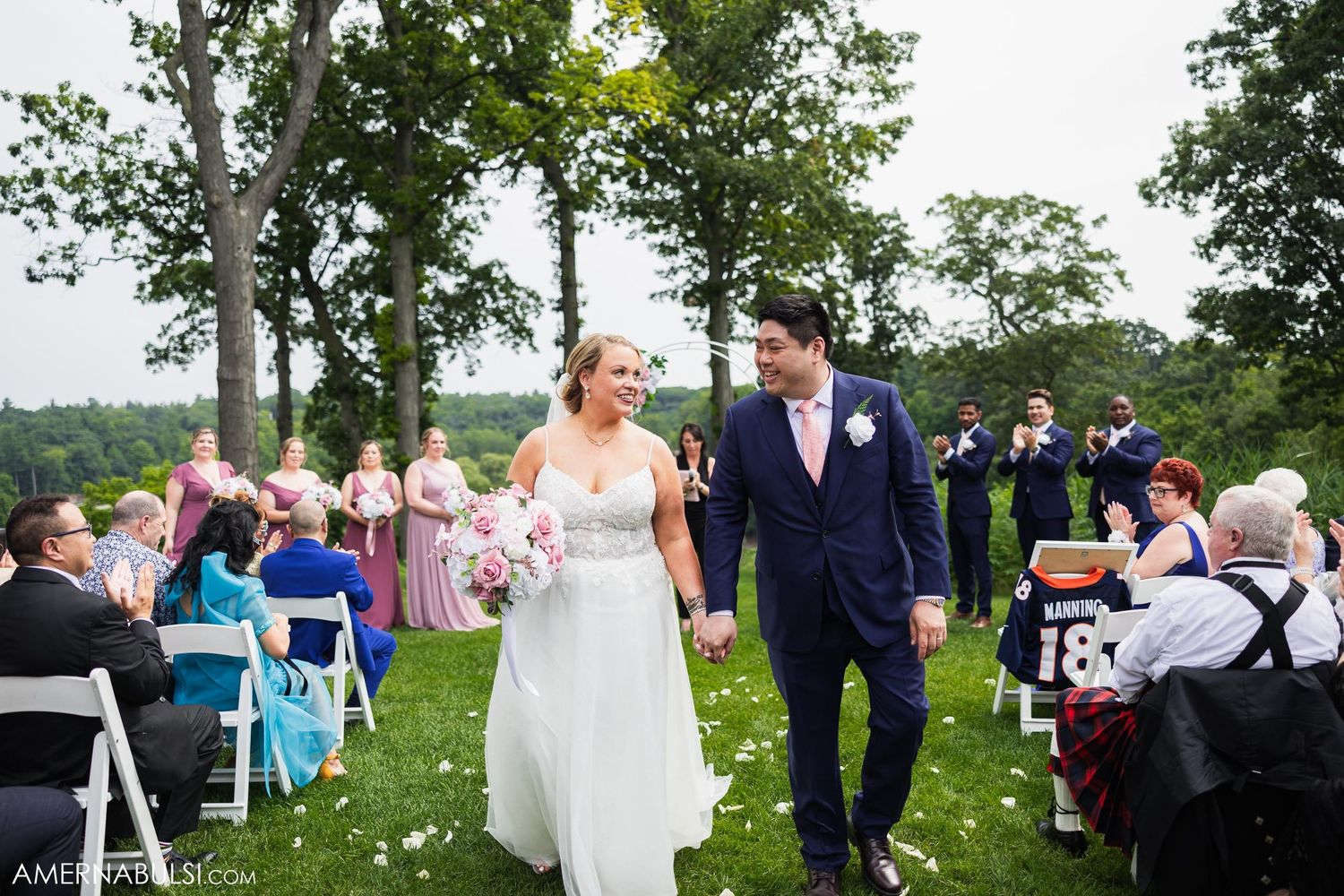 Happy couple walks down the aisle after outdoor ceremony while guests look on from white chairs.