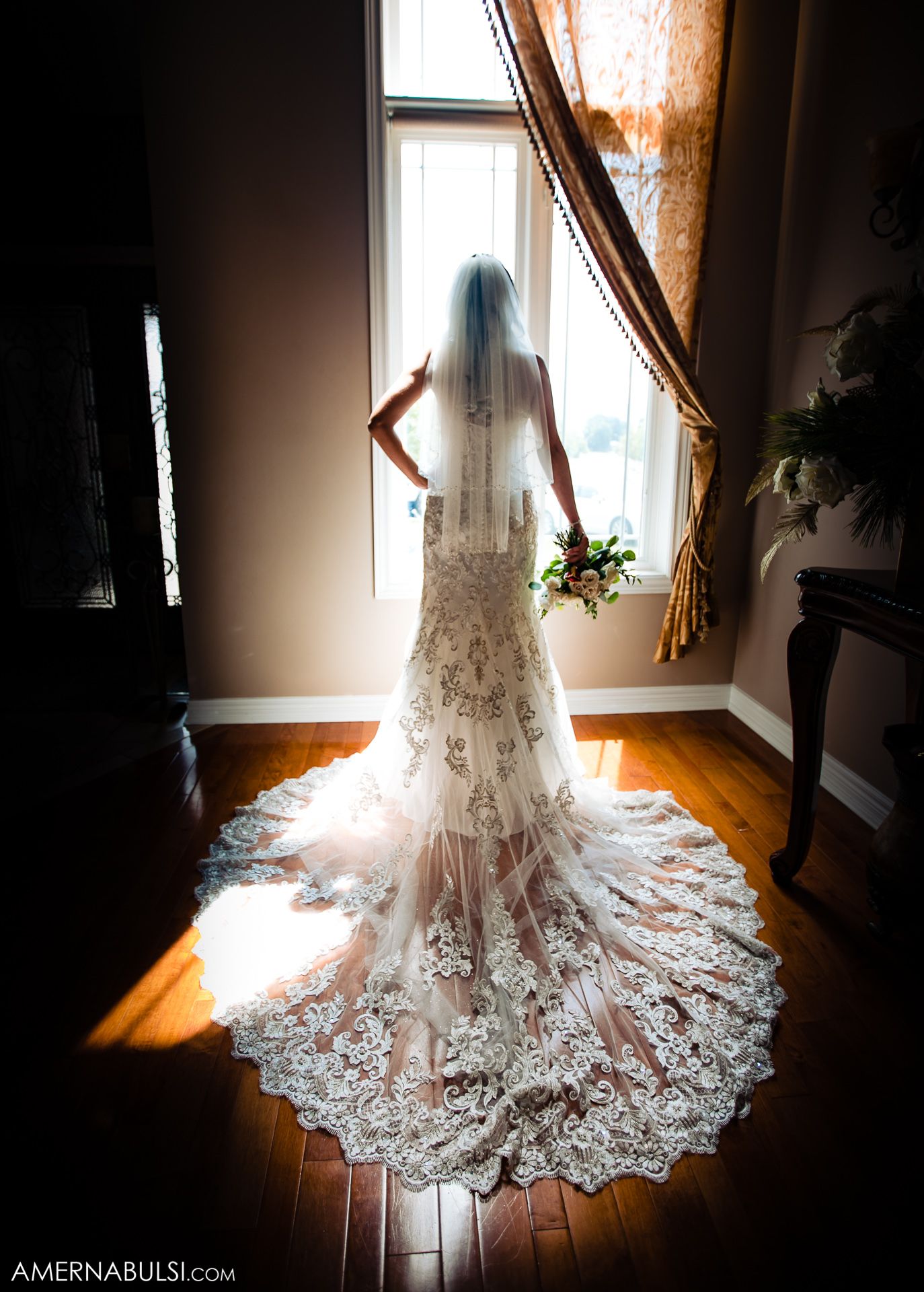 Silhouette of a bride in lace wedding gown with long train standing by window in dramatic lighting.