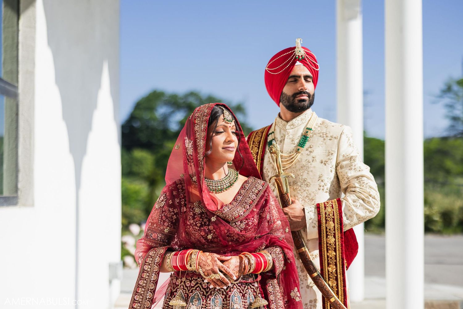 A Sikh couple in traditional wedding attire posing together against white pillars during their wedding ceremony.
