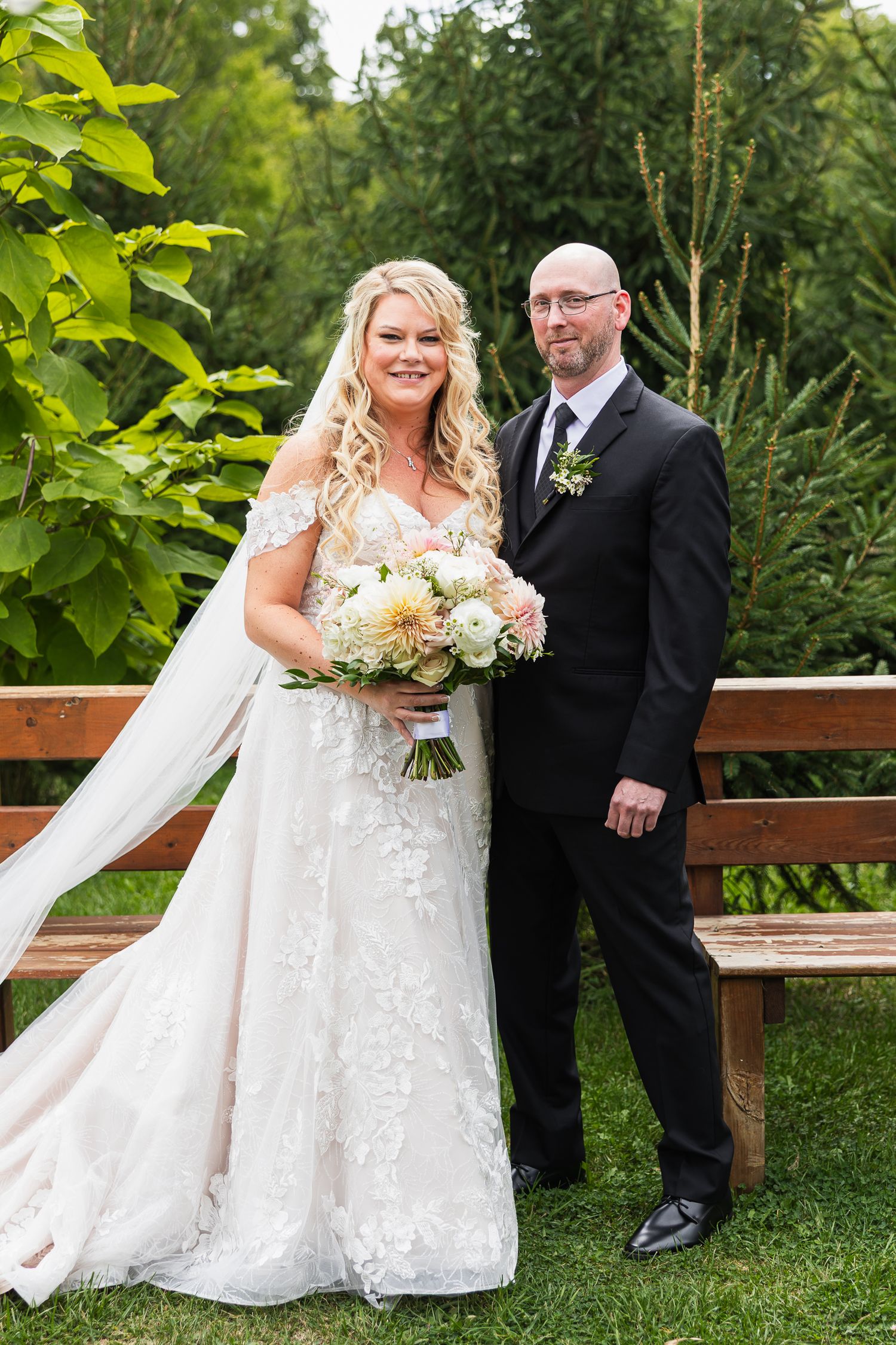 A romantic wedding photoshoot on a wooden bench surrounded by lush green foliage.