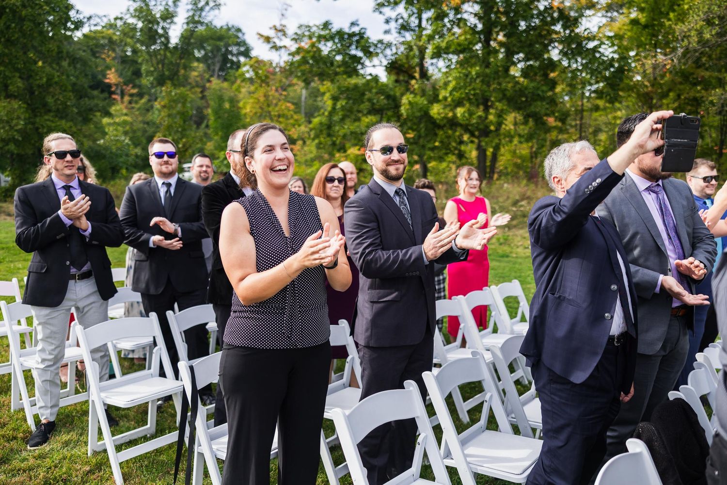 Wedding guests clapping and celebrating during outdoor ceremony with white chairs.