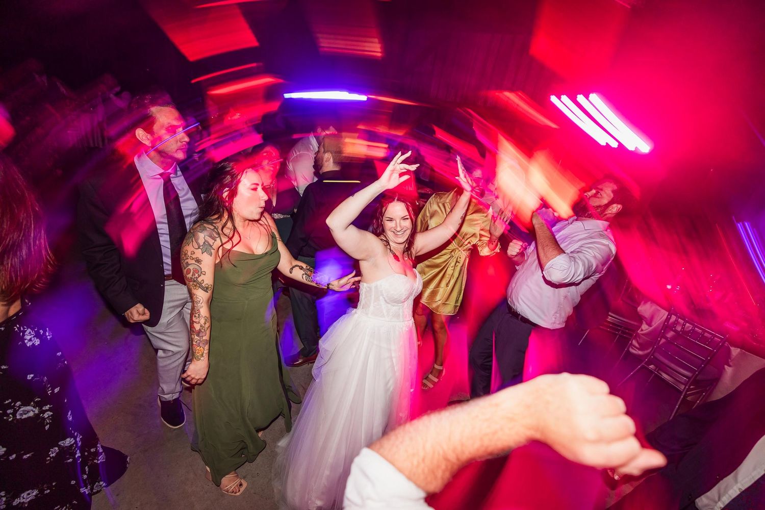 Wedding party dancing energetically under vibrant red and purple lighting at a reception celebration.