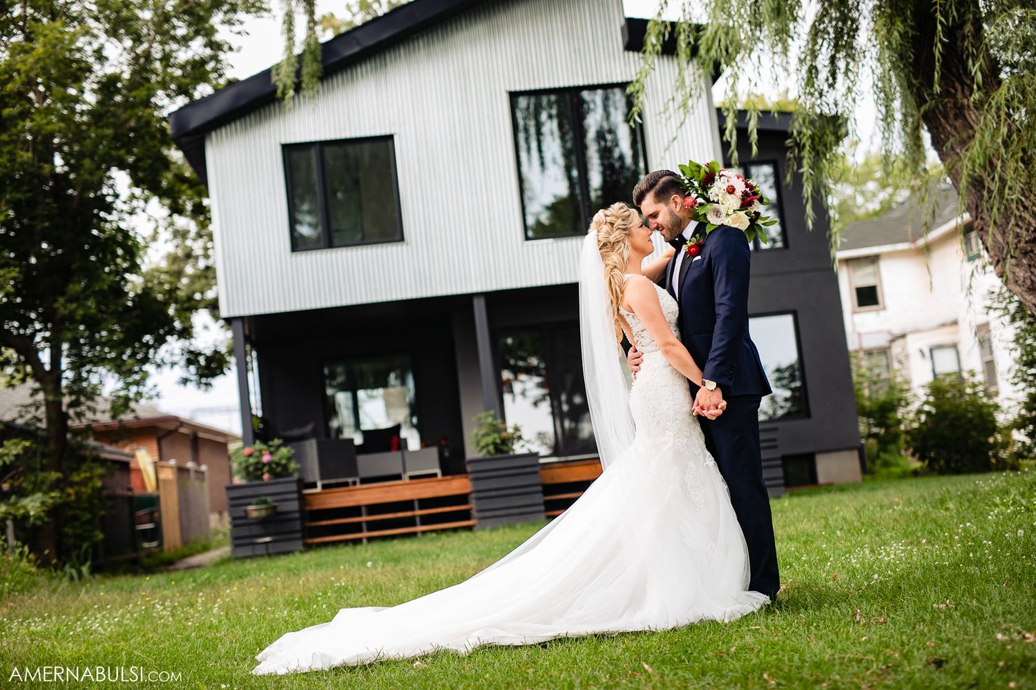 Wedding party poses on lush green lawn in front of white modern building with willow trees.