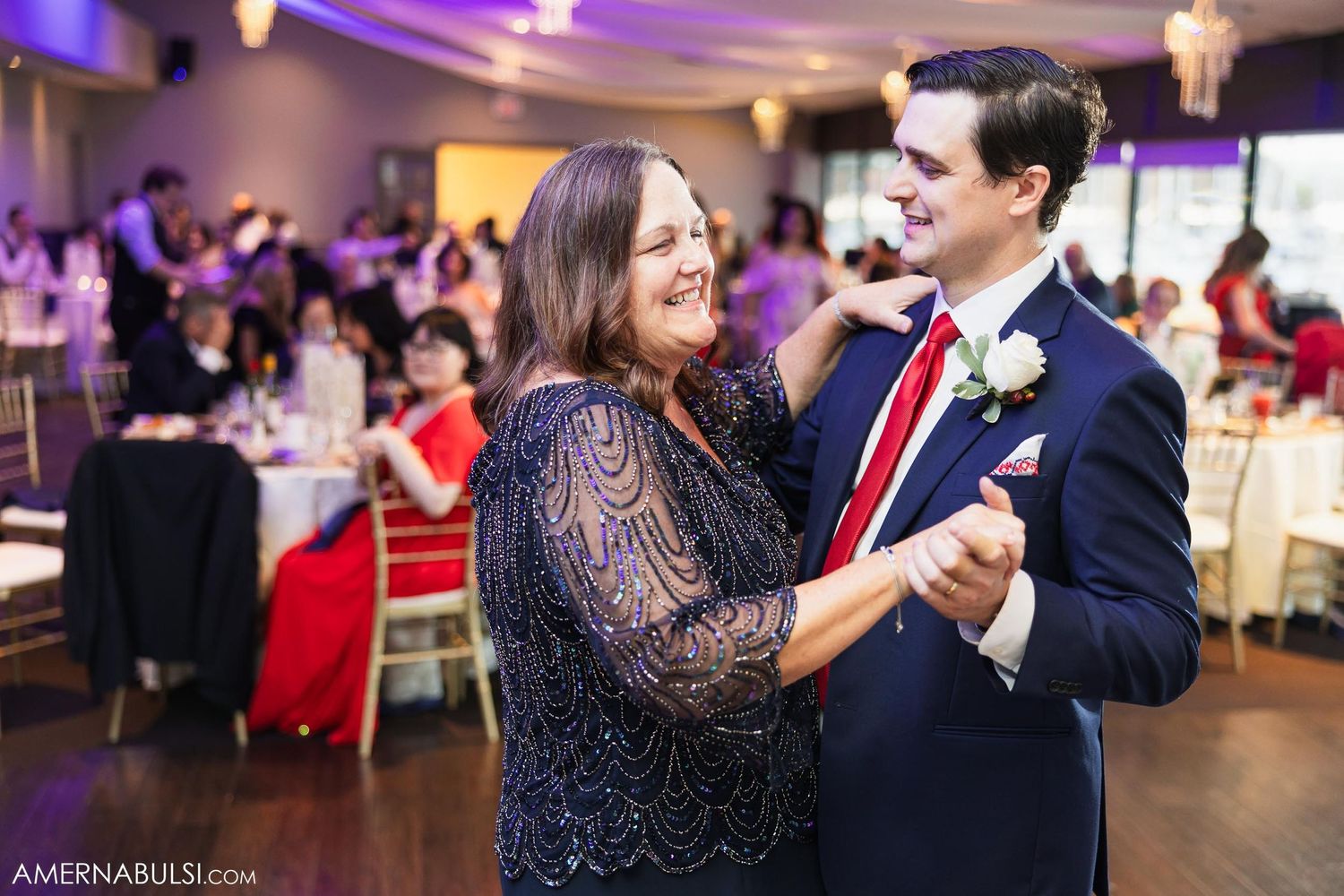 A tender dance moment at a wedding reception with a navy lace dress and red tie against purple lighting.