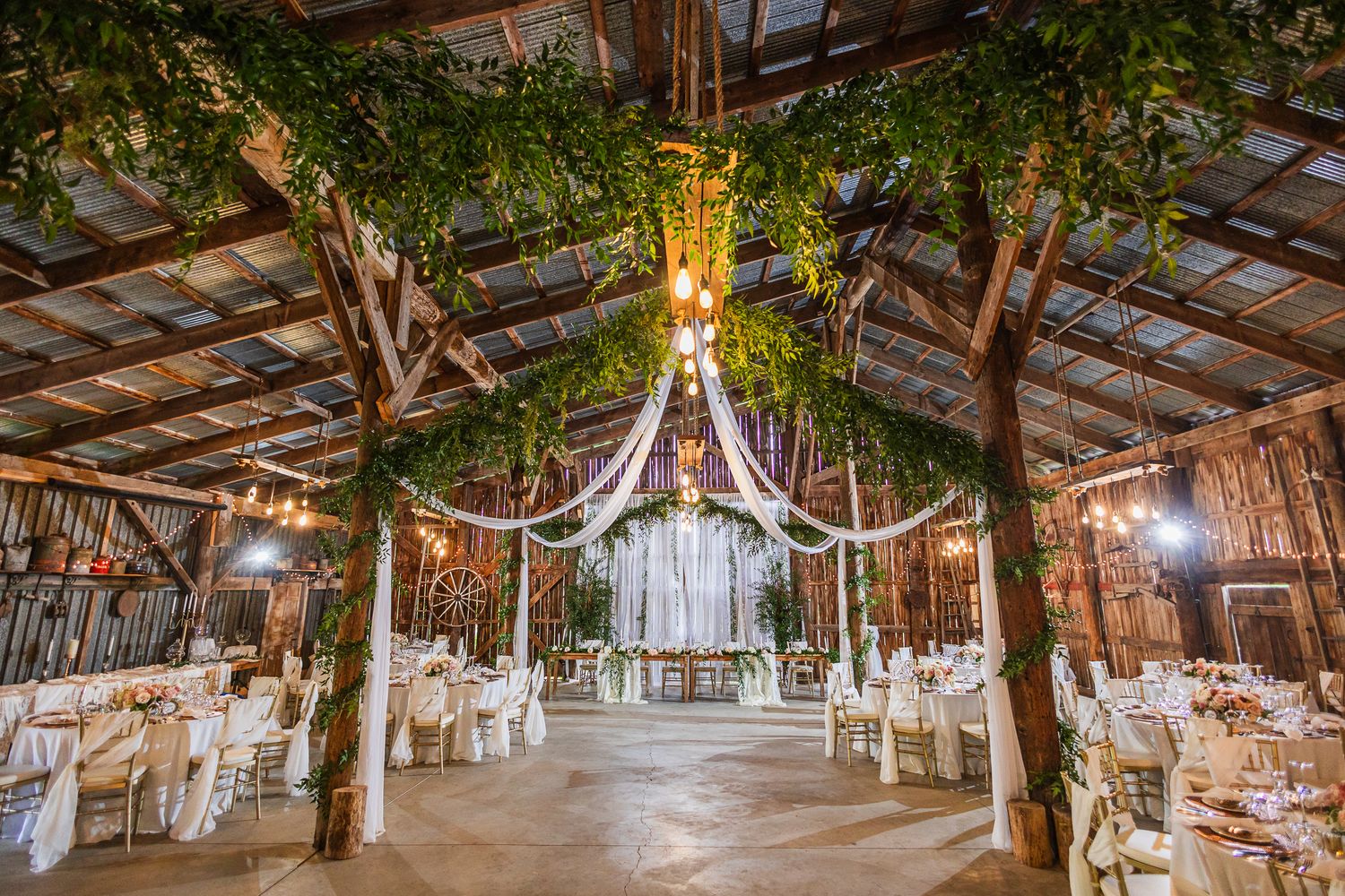 A rustic barn wedding venue interior decorated with string lights and greenery hanging from wooden beams creates an enchanted atmosphere.