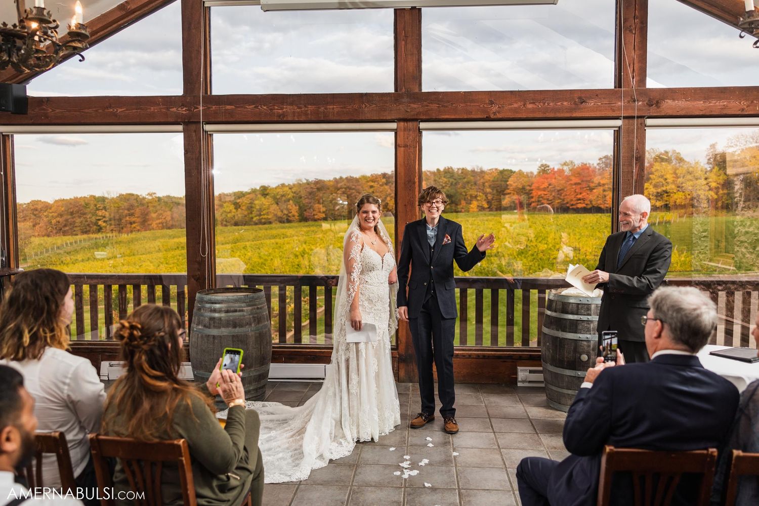 A wedding ceremony in progress with wooden barrel decor and autumn views through large windows.