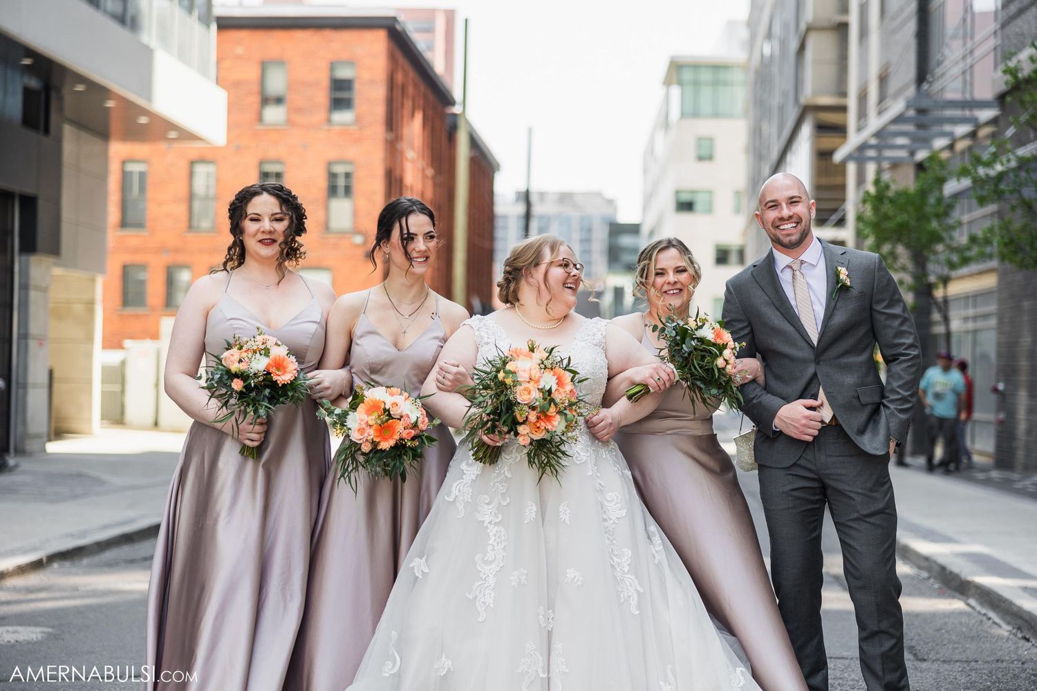 Wedding party poses on urban street wearing mauve dresses and carrying peach bouquets.