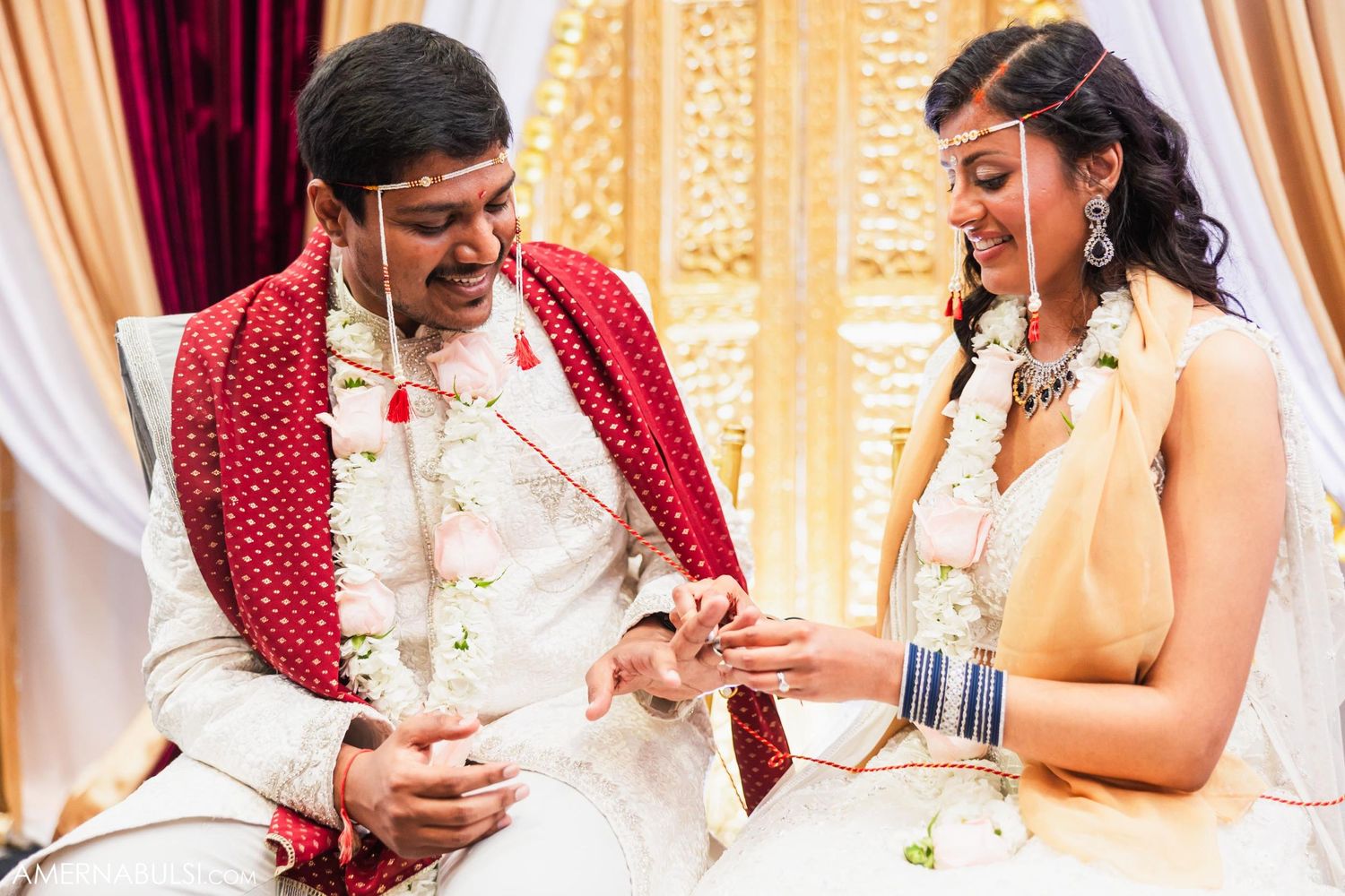 A couple exchange rings during their Indian wedding ceremony wearing traditional white and red attire.