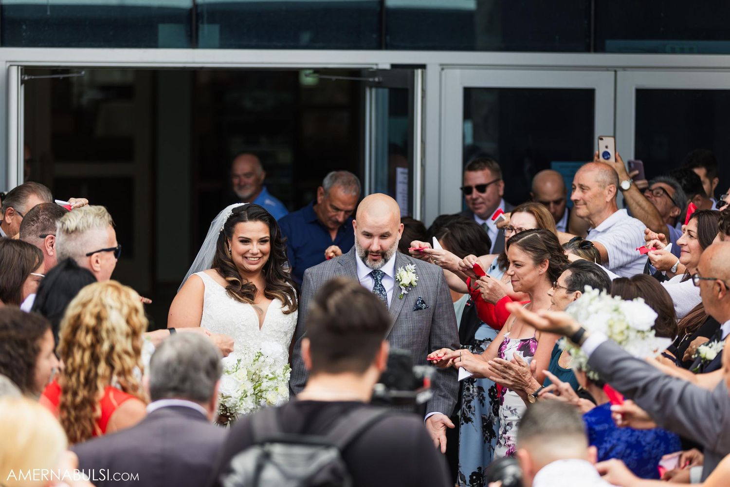 Wedding couple exits building surrounded by celebratory crowd throwing confetti and rose petals.