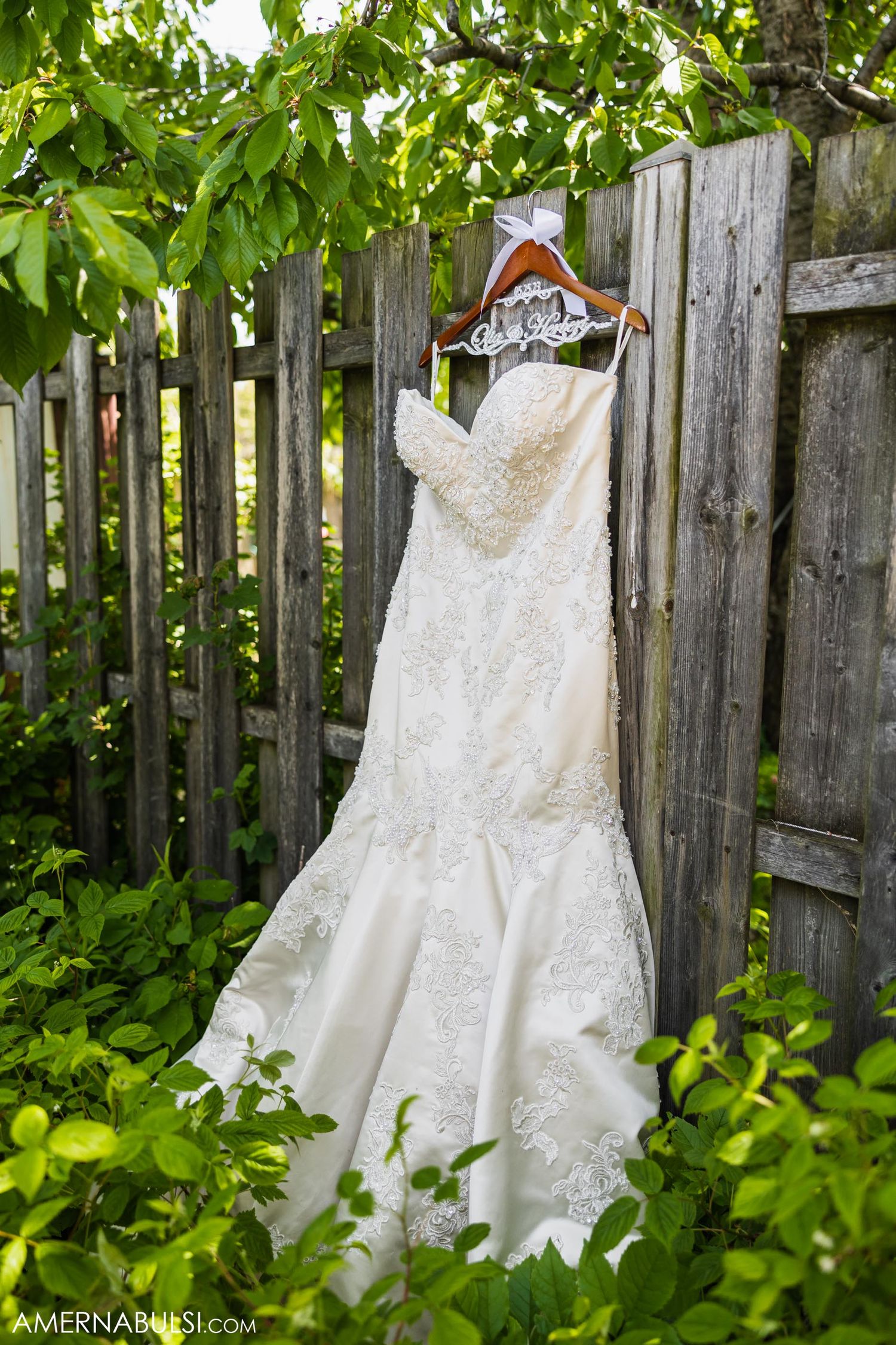 Elegant lace wedding dress hangs on rustic wooden fence surrounded by green foliage.