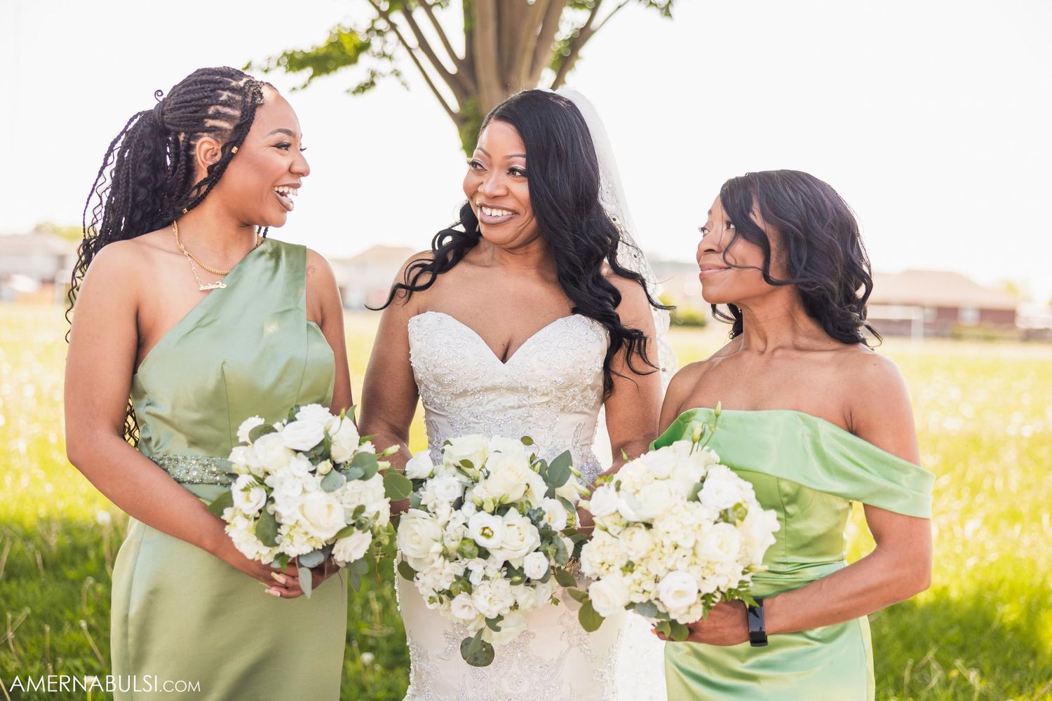 Bridesmaids in sage green dresses laugh with bride holding white bouquets.