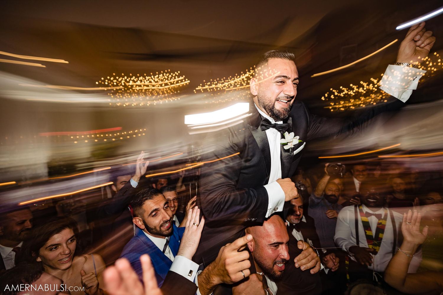 Wedding guests hoist the groom into the air during an energetic reception celebration with twinkling lights overhead.