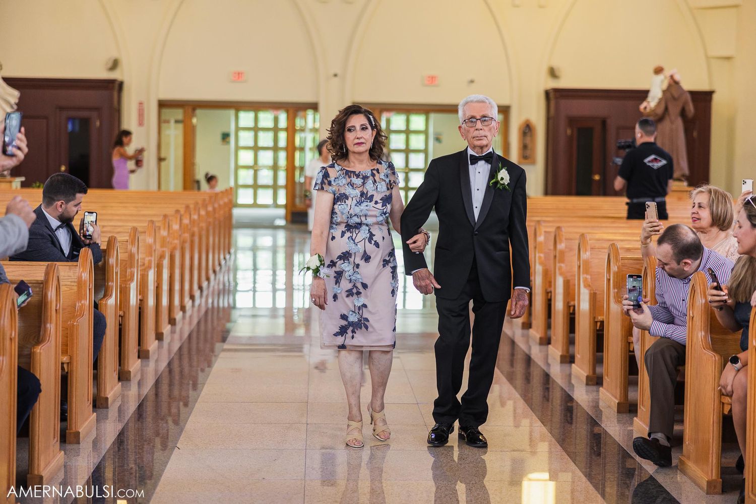 Wedding party members walk down a church aisle while guests capture photos on their phones.