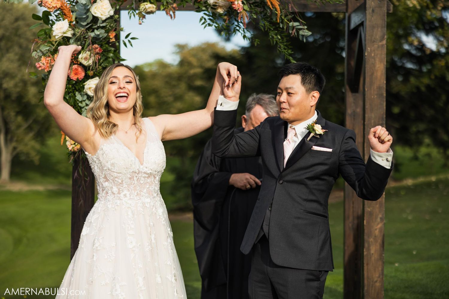 A joyful celebration moment during an outdoor wedding ceremony under a wooden arch with floral decorations.