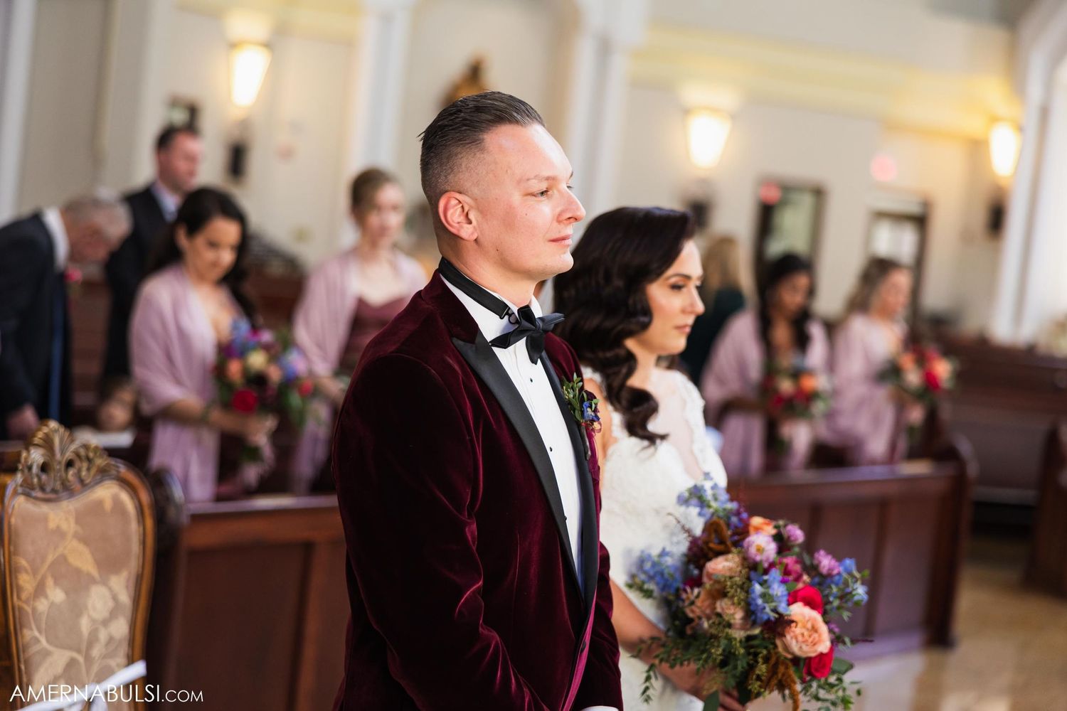Couple during wedding ceremony in church wearing burgundy velvet tuxedo and white gown.