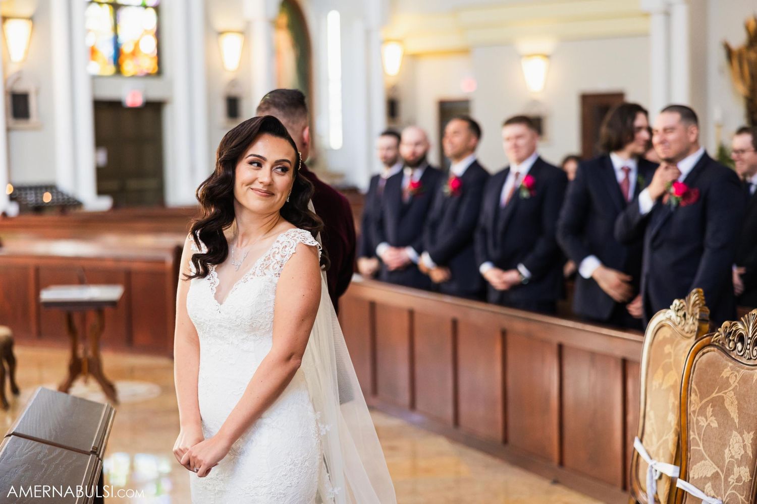A bride in a white lace dress stands at the altar while groomsmen look on from wooden church pews.
