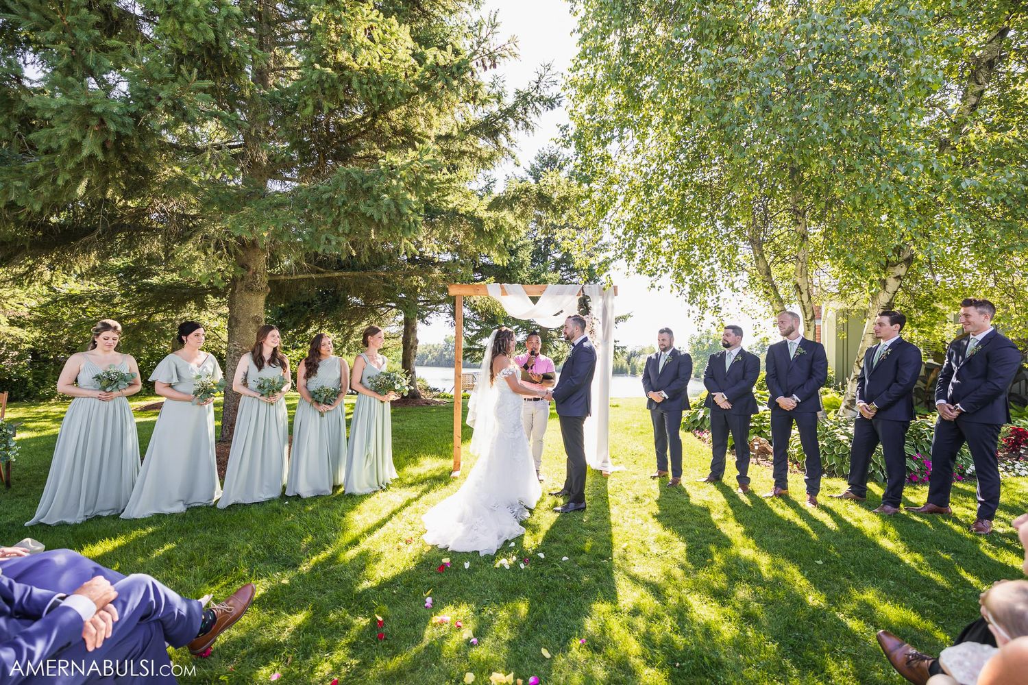 Wedding party arranged on sunny lawn with bridesmaids in light dresses and groomsmen in navy suits.