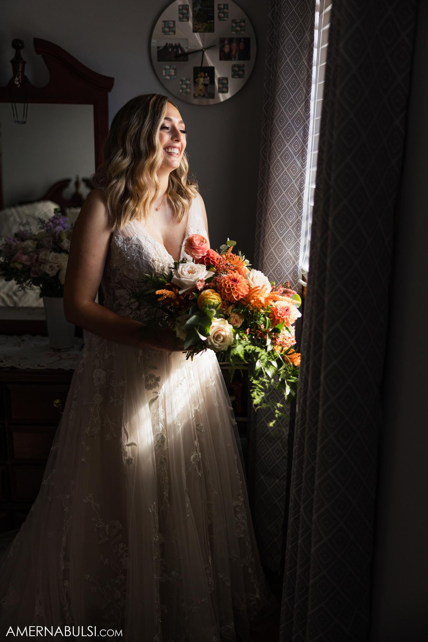 A bride in a lace wedding gown stands by a window holding a vibrant orange and coral bouquet in dramatic lighting.
