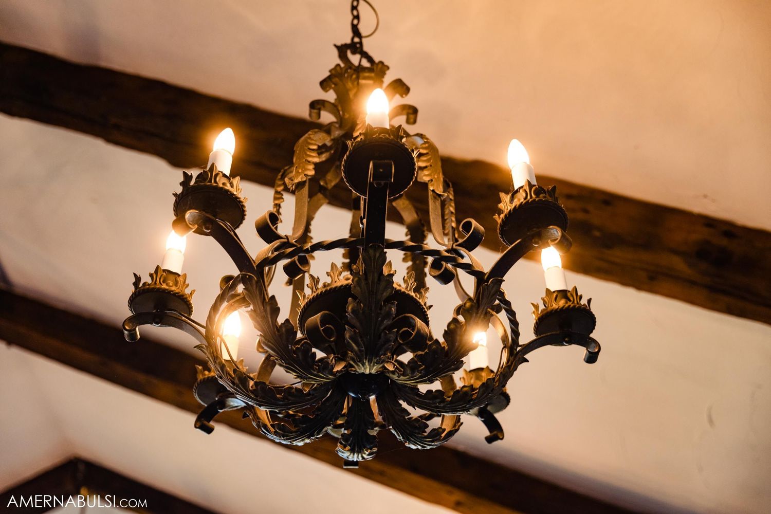 Ornate black iron chandelier with glowing candles hanging from wooden beam ceiling.