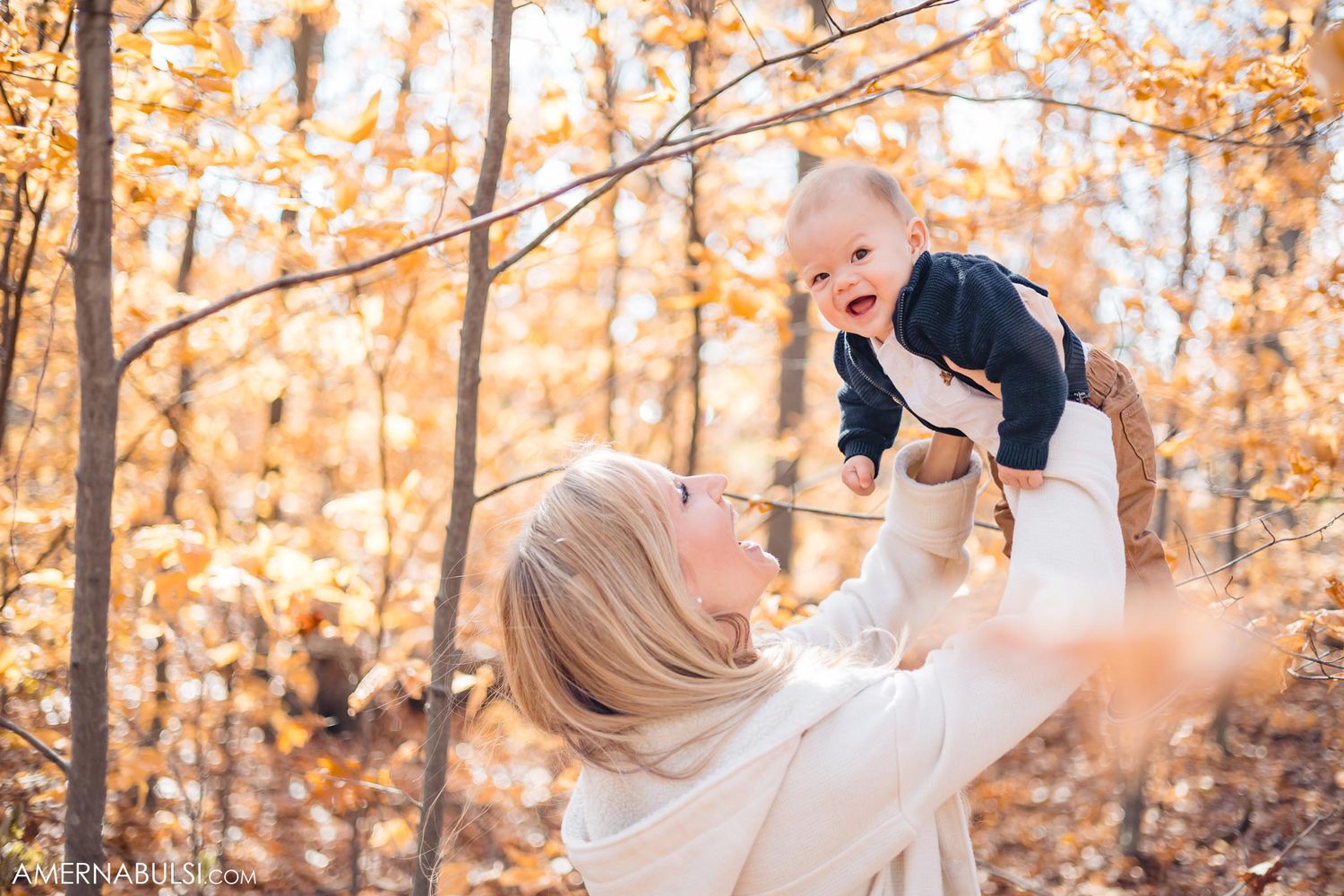 Parent lifts joyful baby in white sweater among golden fall leaves.