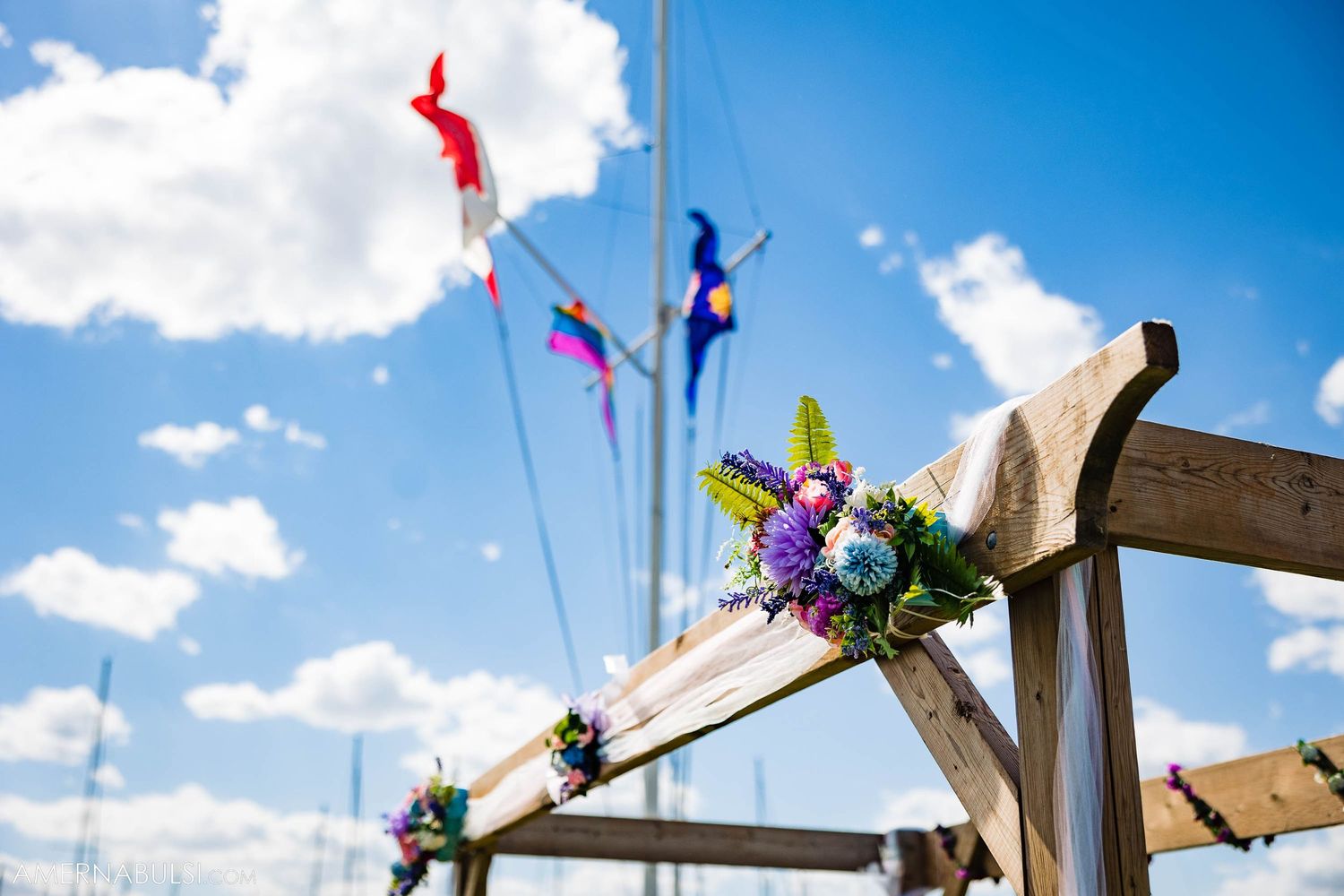 Wooden wedding arch decorated with colorful streamers and purple flowers against blue sky.