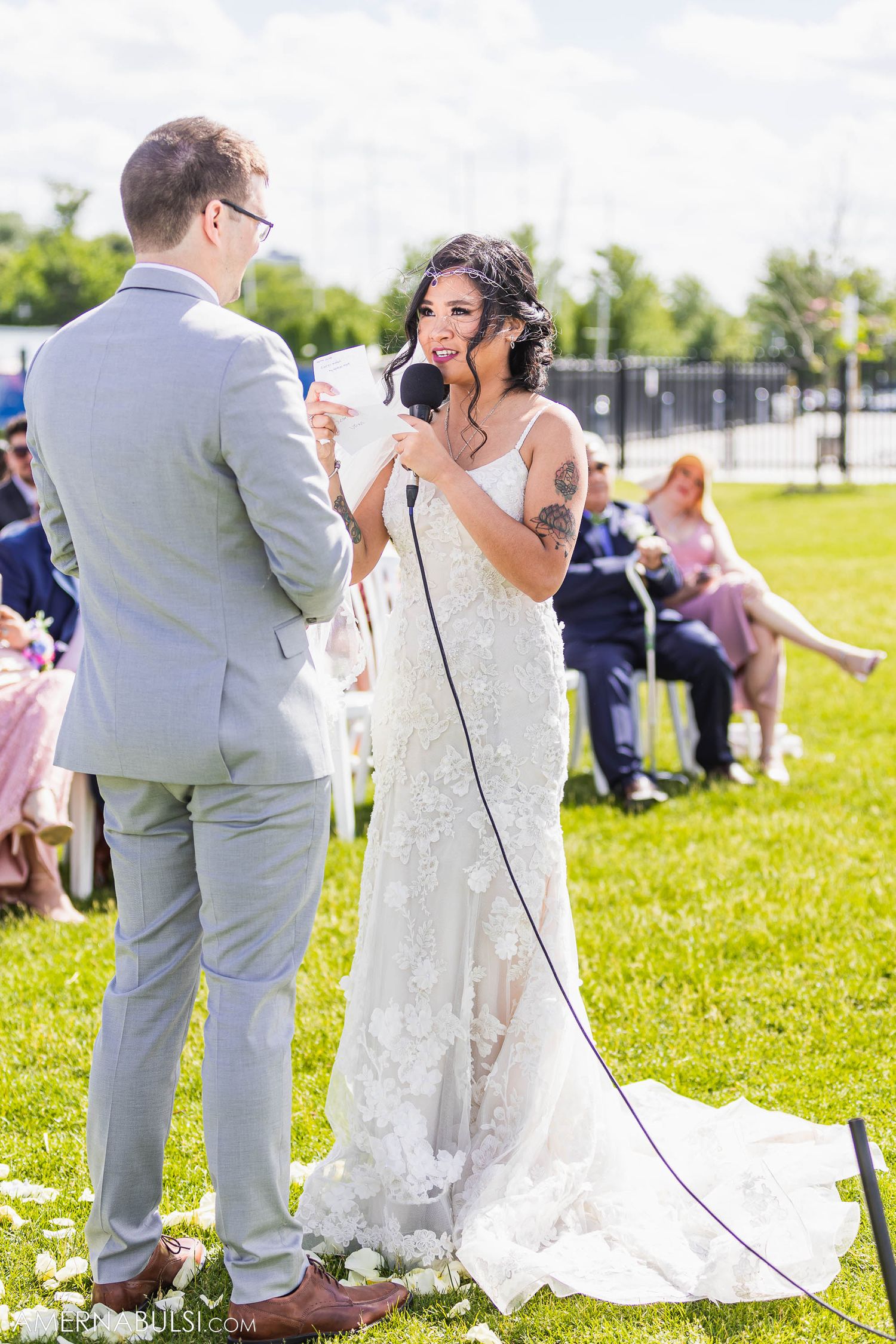 Couple shares wedding vows on green lawn with guests seated in background.