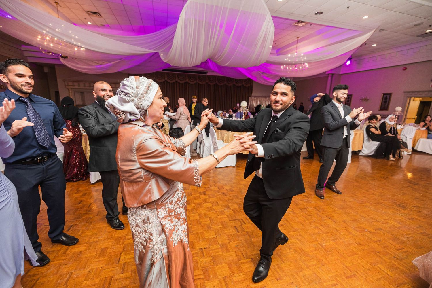 People dancing energetically on a wooden dance floor at a wedding reception with purple ambient lighting.