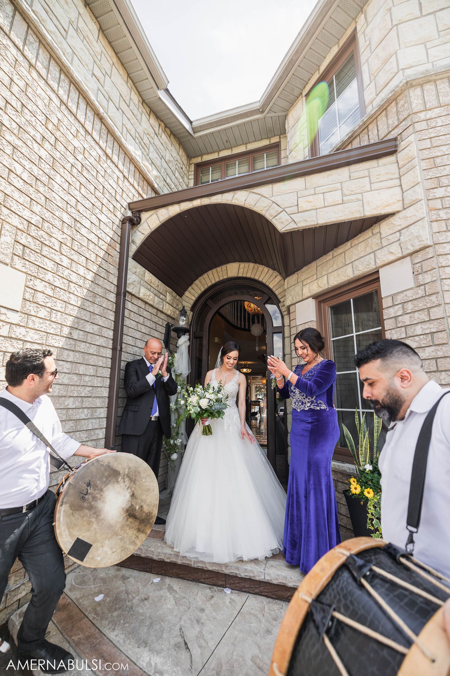 Wedding party exits stone building entrance accompanied by musicians with drums.