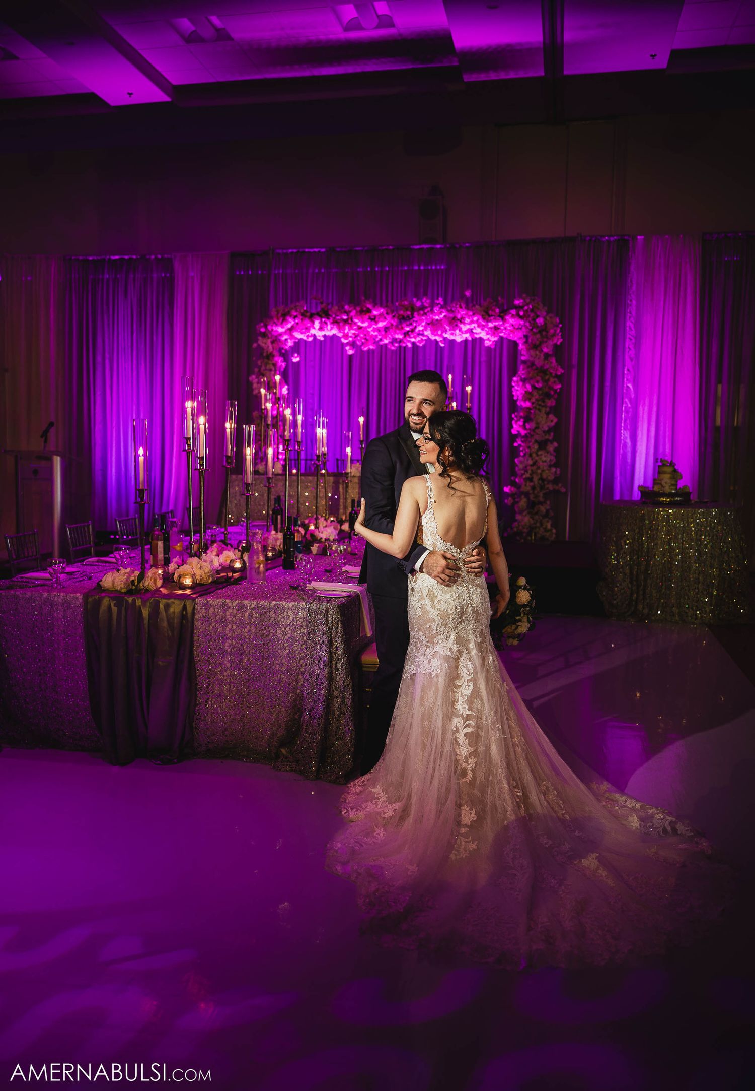 Romantic reception portrait of newlyweds under purple lighting with floral arch backdrop.